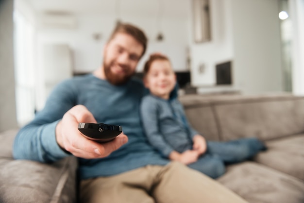 Foto van een vrolijke, bebaarde vader die de afstandsbediening vasthoudt terwijl hij tv kijkt met zijn schattige zoontje. Focus op afstandsbediening.
