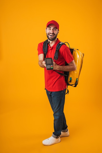 Foto van een vrolijke bezorger in een rood uniform met een betaalterminal terwijl hij een rugzak draagt met afhaalmaaltijden geïsoleerd over geel