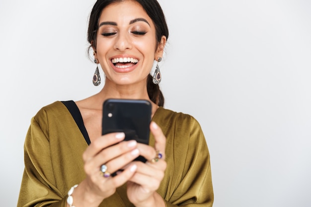 Foto van een vrolijke Indiase vrouw met make-up die etnische sieraden draagt en een traditionele zijden jurk die poseert met een mobiele telefoon geïsoleerd over een witte muur
