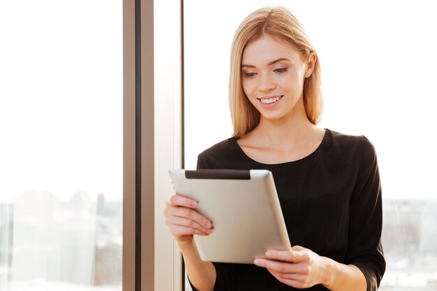 Foto van een vrolijke jonge dame die in kantoor staat met een tabletcomputer. Kijk naar tabletcomputer.