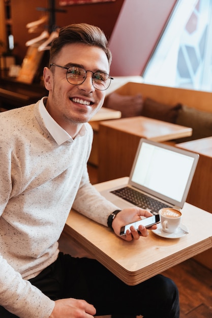 Foto van een vrolijke jonge man met een bril die in café in de buurt van koffie en laptop zit terwijl hij smartphone gebruikt. Camera kijken.