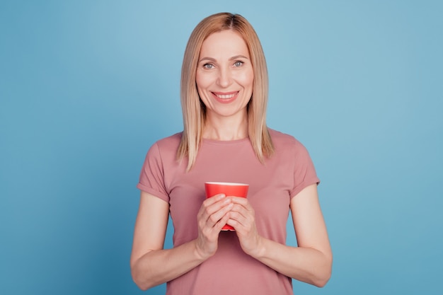 Foto van een vrolijke positieve vrouw die een mok koffie vasthoudt die tandjes glimlacht, geïsoleerd over een blauwe kleurachtergrond