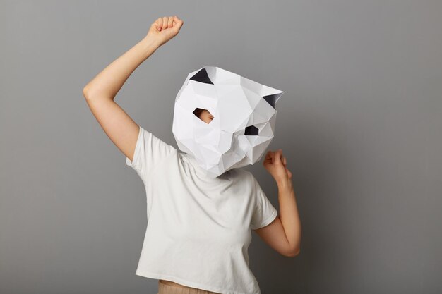 Foto van een vrolijke, vrolijke anonieme vrouw met een kattenmasker en een wit t-shirt poseren met opgeheven armen dansen op een themafeest geïsoleerd over grijze achtergrond