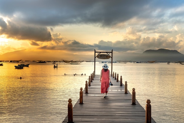 Foto van een vrouw in een roze outfit en een zonnehoed die op een houten kade over het meer loopt terwijl ze iets draagt