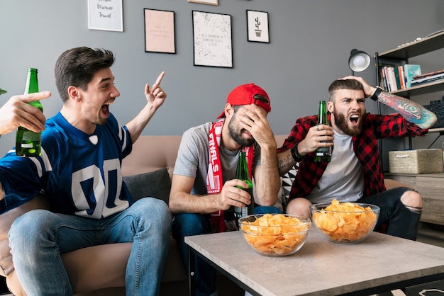 Foto van fans van blanke emotionele jongens die schreeuwen en bier drinken tijdens het kijken naar sportwedstrijden in de woonkamer