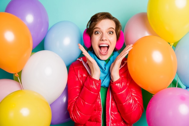 Foto van gekke dame omringd door vele kleurrijke luchtballonnen verrassing verjaardagsfeestje verheugt zich dragen casual rode jas sjaal trui roze oorkappen geïsoleerde groenblauw kleur muur