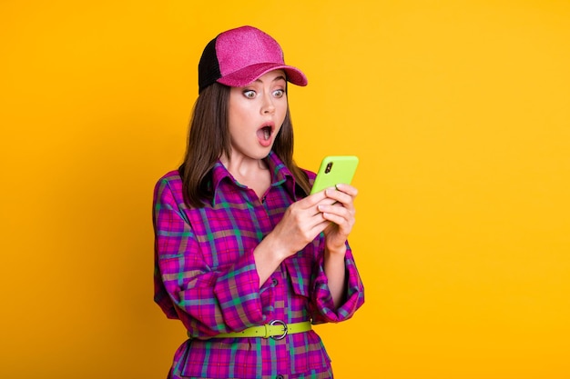 Foto van geschokt mooi meisje dat smartphone met open mond magenta hoofddeksels staart geïsoleerd op gele kleur achtergrond