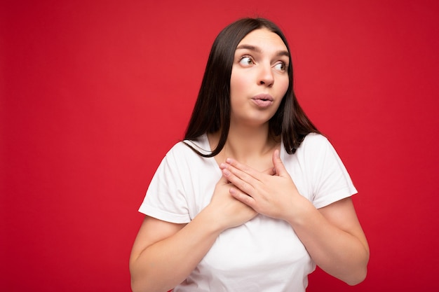 Foto van geschokte verbaasde treurige jonge mooie brunette vrouwelijke persoon met oprechte emoties die casual wit t-shirt draagt voor mockup geïsoleerd op rode achtergrond met kopieerruimte