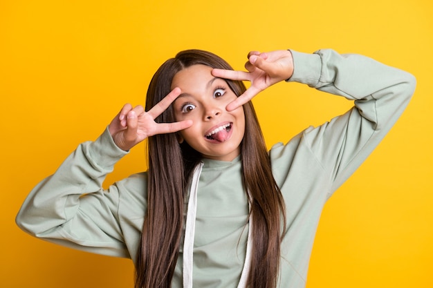 Foto van grappig lief schoolmeisje draagt een casual grijze outfit met v-teken ogen tong geïsoleerde gele kleur achtergrond