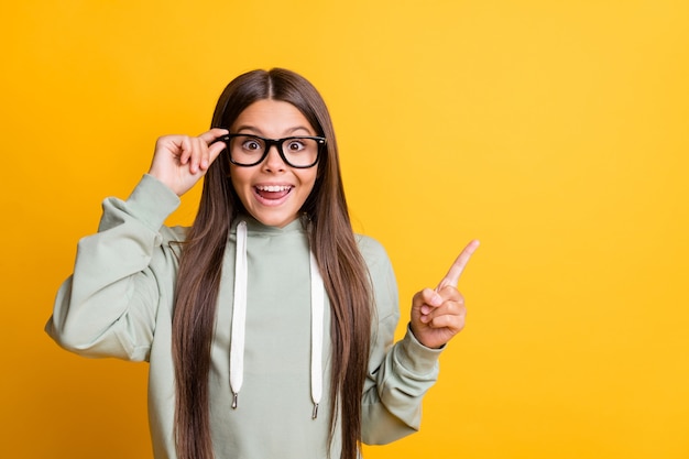 Foto van grappig schattig schoolmeisje draagt een casual grijze outfit arm hand bril wijzend op lege ruimte geïsoleerde gele kleur achtergrond