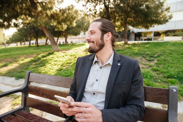 Foto van harige bebaarde zakenman 30s mobiele telefoon houden en opzij kijken, terwijl u geniet van rust in het stadspark