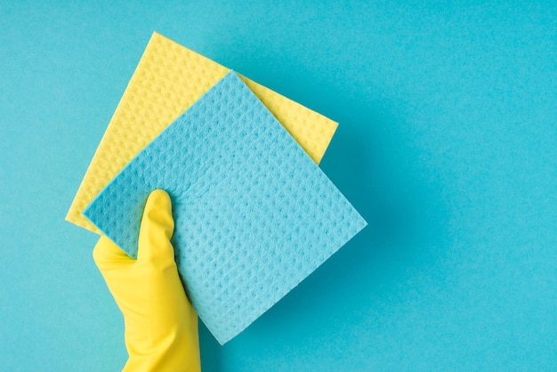 Foto van het bovenaanzicht van de hand in gele rubberen handschoen met gele en blauwe viscosevodden op geïsoleerde pastelblauwe achtergrond met copyspace