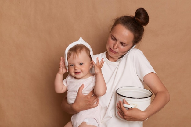 Foto van huilende moeder die een wit T-shirt draagt en haar peuterdochter in handen houdt en in de pot kookt met baby-baby pratende telefoon en gestresst poseren geïsoleerd op bruine achtergrond