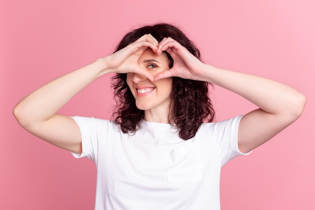 Foto van jong mooi meisje met vingers, hartfiguur, kijk valentijnsdag geïsoleerd over roze kleurachtergrond