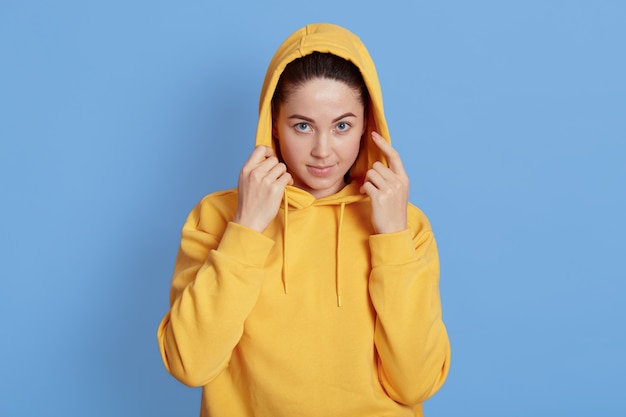 Foto van jonge donkerharige vrouw met natuurlijke make-up hand in hand op haar kap terwijl ze aandachtig naar de camera kijkt, staande op blauwe achtergrond, jurken gele hoodie.