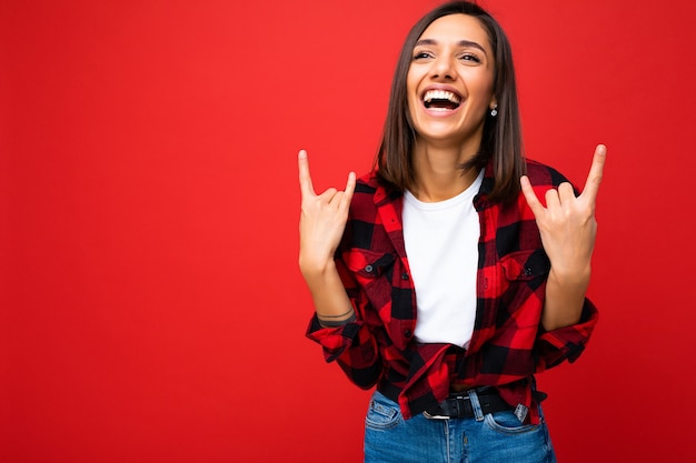 Foto van jonge gelukkig positieve coole aantrekkelijke brunette vrouw met oprechte emoties, gekleed in wit t-shirt en hipster rood geruit overhemd geïsoleerd op rode muur met lege ruimte en plezier.