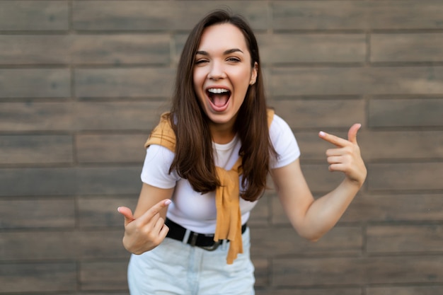 Foto van jonge gelukkig vreugdevolle aantrekkelijke brunette vrouw met oprechte emoties stijlvolle outfit dragen staande in de straat in de buurt van bruine muur en wijzend op zichzelf. Levensstijl concept.