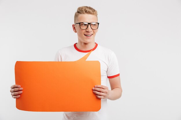 Foto van jonge Kaukasische kerel die vrijetijdskleding dragen en glazen die copyspace oranje affiche in handen voor uw tekst houden op wit wordt geïsoleerd