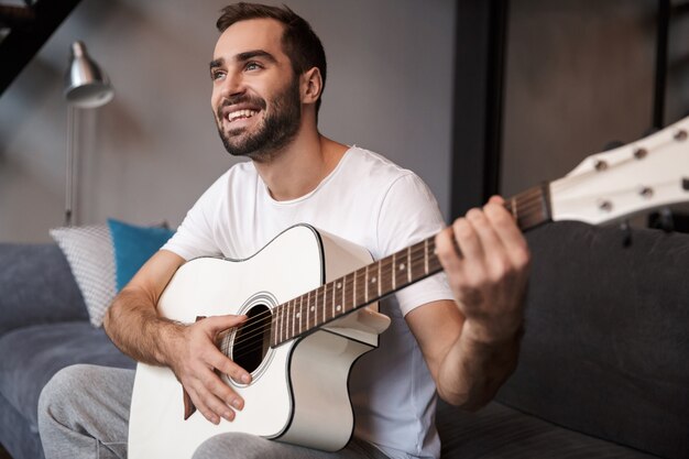 Foto van jonge man 30s dragen casual t-shirt akoestische gitaar spelen zittend op de Bank in appartement