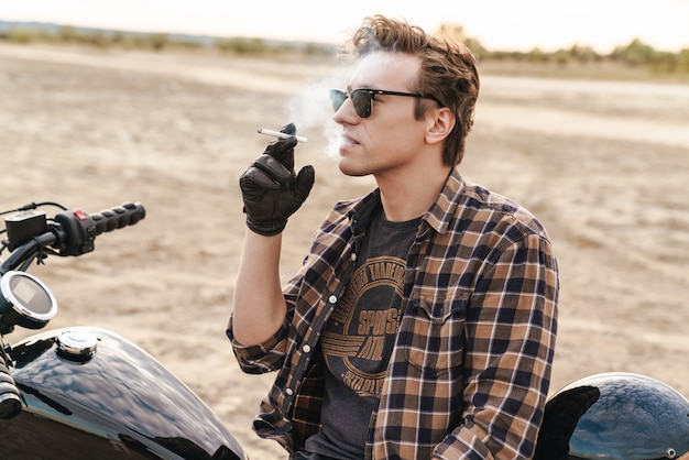 Foto van jonge man biker buiten op het woestijnveld roken sigaret.