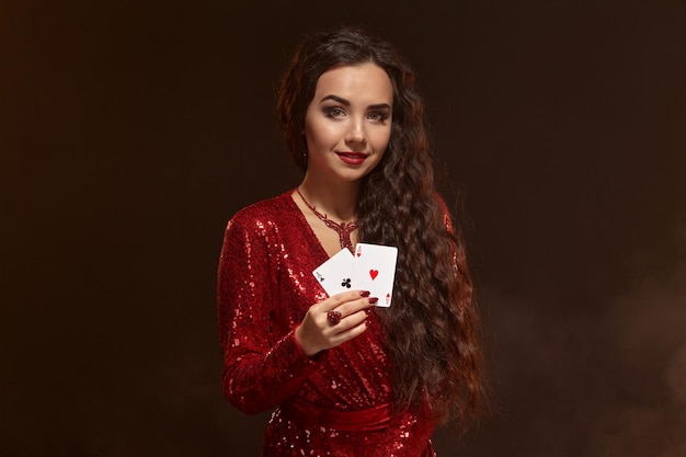 Foto van jonge mooie blanke brunette vrouw in glanzende rode avondjurk houdt kaarten in één hand, toont een paar azen. bruine achtergrond, casino, gokindustrie