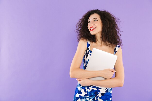 Foto van jonge mooie vrouw met krullend haar in kleding die en laptop in handen glimlacht houdt