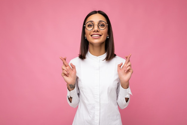 Foto van jonge positieve gelukkig lachende mooie vrouw met oprechte emoties, gekleed in stijlvolle kleding geïsoleerd op de achtergrond met kopieerruimte en met vingers gekruist voor geluk.