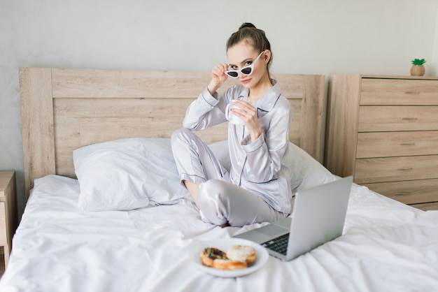 Foto van jonge vrolijke vrouw in pyjama typen op laptop en glimlachen zittend op bed in de kamer