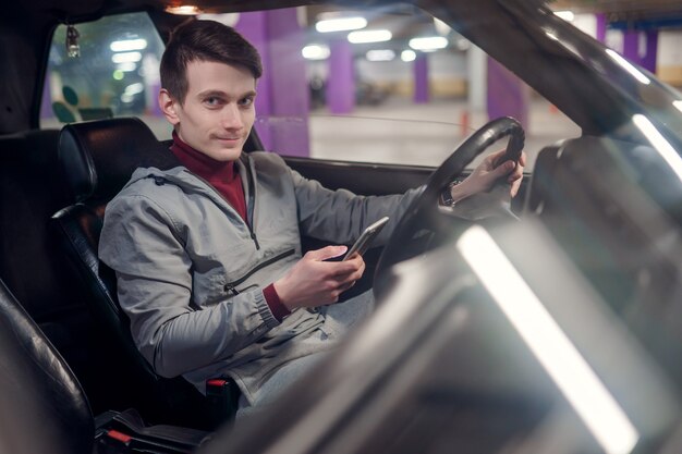 Foto van kant van lachende mannelijke chauffeur met telefoon in handen zittend in auto op ondergrondse verzekering