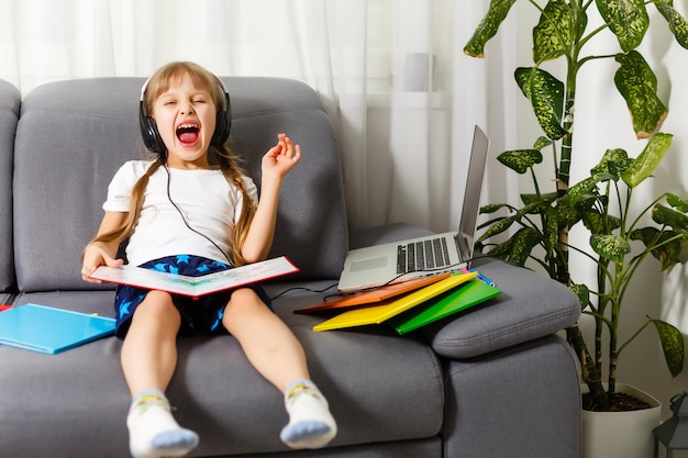 Foto van kleine mooie leerling school dame zet skype laptop notitieboekje online les open houd boek klaar antwoord leraar vragen zit bank afstand quarantaine studie comfort kamer binnenshuis