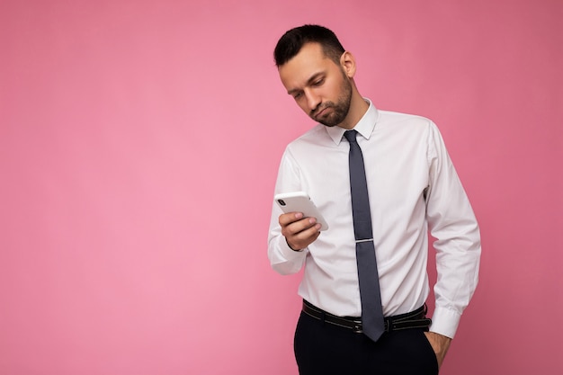 Foto van knappe knappe man met casual wit overhemd en stropdas geïsoleerd op roze achtergrond met lege ruimte in de hand houden en met behulp van mobiele telefoon messaging sms camera kijken. kopieer ruimte