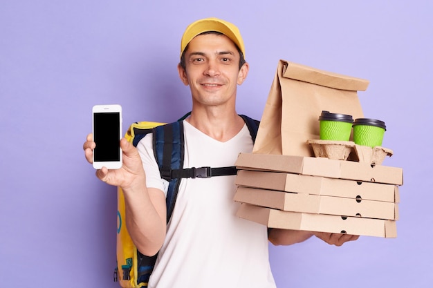 Foto van lachende aantrekkelijke man koerier in gele dop en t-shirt met eten bestellen en smartphone met lege display die pizzadozen en koffie brengt om naar de klant te gaan geïsoleerd op paarse achtergrond