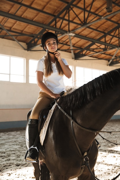 Foto van lachende tevreden jonge blonde mooie vrouw met hoed met paard op het platteland in corral op trainingsgebied.