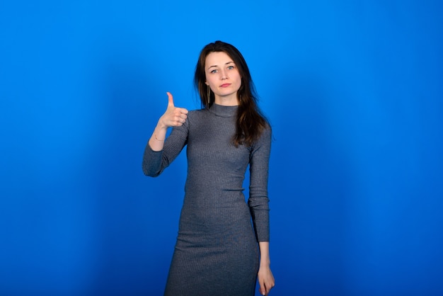 Foto van lachende, vrolijke jonge vrouw in grijze jurk op blauwe muur. Emotioneel portret