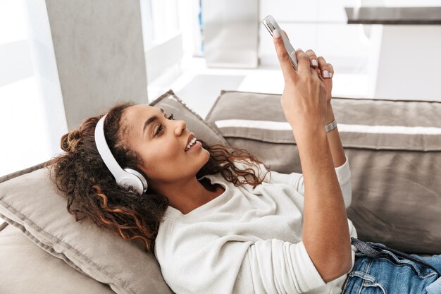 Foto van moderne afrikaanse amerikaanse vrouw die hoofdtelefoons draagt die celtelefoon houden, terwijl liggend op bank in helder appartement