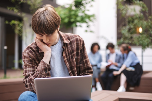 Foto van moe jongeman zittend op een bankje met laptop op knieën en denken terwijl tijd doorbrengen op de binnenplaats van de universiteit met studenten op de achtergrond