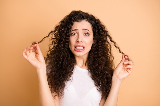 Foto van mooie dame met golvende permanente krullen in slechte conditie na een bezoek aan stylist paniek gespleten haarpunten dragen witte vrijetijdskleding geïsoleerde beige pastelkleur achtergrond