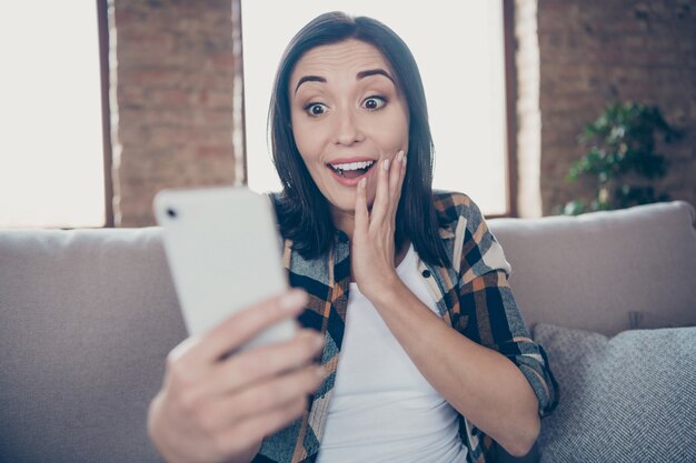 Foto van mooie dame met telefoon lezen van nieuwe instagram post positieve reacties dolblij zitten gezellige bank dragen casual kleding appartement binnenshuis