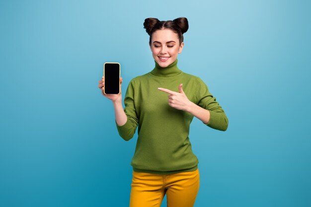 Foto van mooie dame verkoopmanager houden nieuw model smartphone tonen huidige advertentie verbazingwekkende kwaliteit apparaat dragen groene coltrui gele broek geïsoleerde blauwe kleur muur