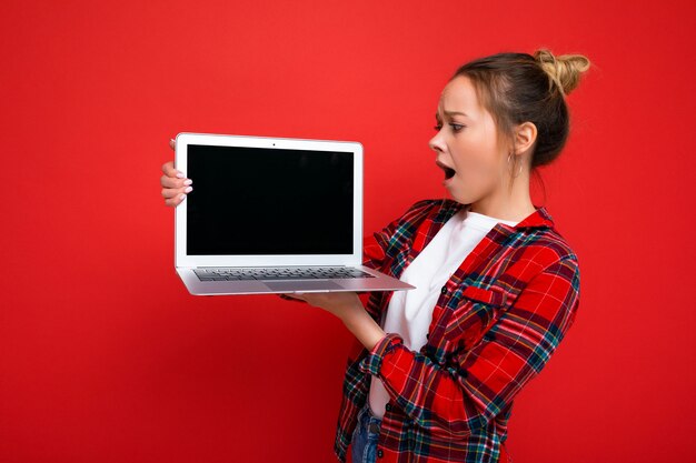 Foto van mooie geschokt verbaasde jonge vrouw met laptop draagt â € ‹â €‹ een rood shirt kijken naar monitorweergave geïsoleerd op rode achtergrond. Bespotten