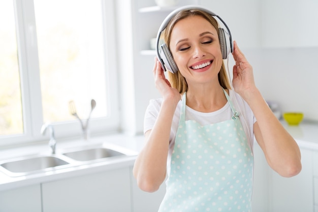 Foto van mooie huisvrouw besteden weekendochtend luisteren naar muziek met behulp van coole moderne oortelefoons plezierige melodie energie krijgen voor algemene schoonmaak witte keuken binnenshuis