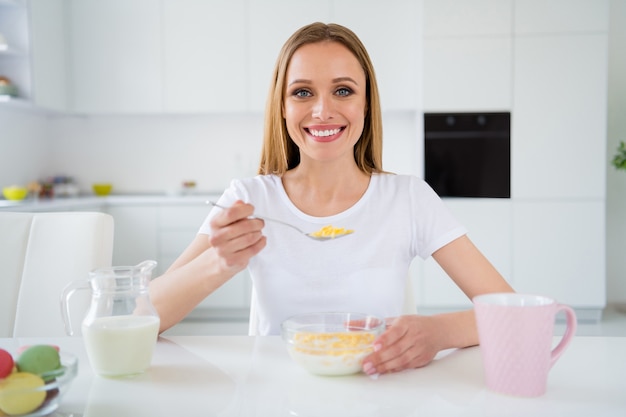 Foto van mooie huisvrouw die volledige lepel verse melk ontbijt cornflakes houdt lekker gezond eten geniet van lekkere maaltijd zittend tafel wit licht keuken binnenshuis