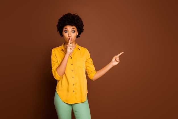 Foto van mooie mooie donkere huid dame wijzend op vingers lege ruimte vinger op lippen adviseren verkoop winkelen prijzen dragen geel shirt groene broek geïsoleerde bruine kleur