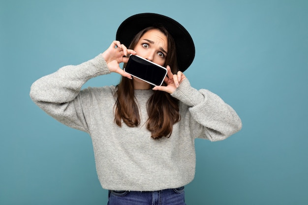 Foto van mooie positieve vrouwenpersoon die zwarte hoed en grijze sweater draagt die mobilofoon houdt