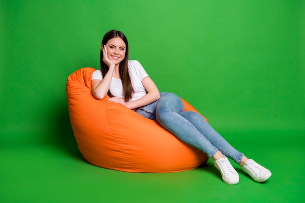 Foto van mooie schattige brunette haar jonge vrouw hand wang jurk wit t-shirt jeans schoeisel zitten op poef geïsoleerd op groene achtergrond