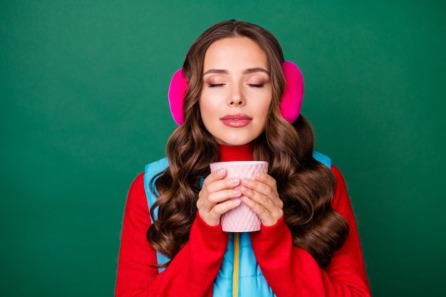 Foto van mooie schattige dromerige jonge dame houd kopje koffie drinken dichte ogen ruiken perfecte smaak draag roze oorwarmers blauw vest met rits rode trui geïsoleerde groene kleur achtergrond