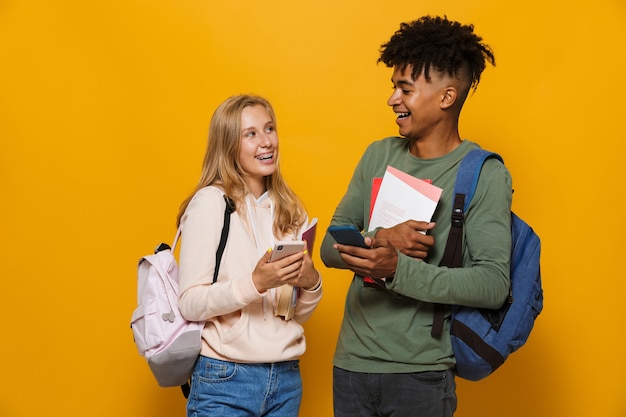 Foto van mooie studenten, man en vrouw 16-18 met rugzakken die glimlachen terwijl ze oefenboeken vasthouden en mobiele telefoon gebruiken, geïsoleerd op gele achtergrond