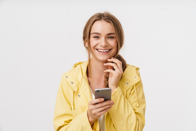 Foto van mooie vrolijke vrouw in regenjas die mobiel gebruikt en glimlacht