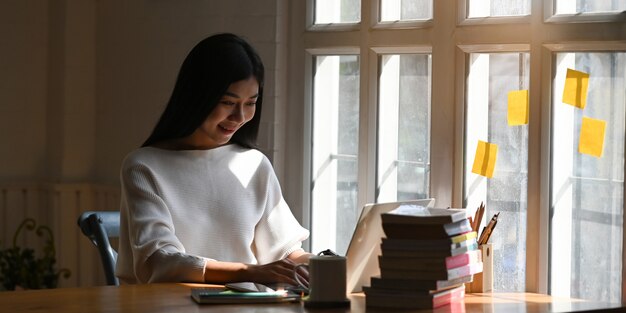 Foto van mooie vrouw die een smartphone in haar handen houden terwijl het zitten bij de houten lijst vooraan computerlaptop, koffiekop, stapel boeken en notitieboekje in ordelijke bibliotheek.