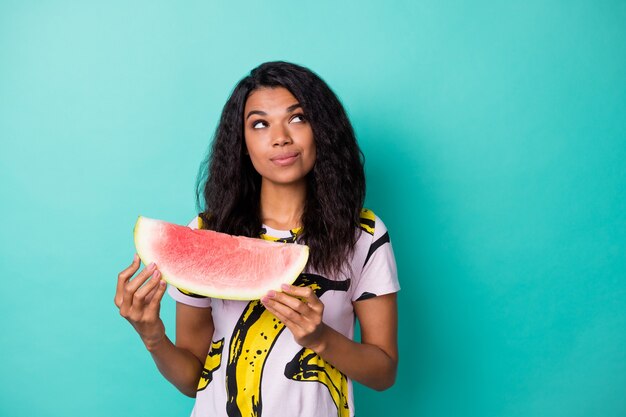 Foto van nieuwsgierig meisje met donkere huid ziet er lege ruimte uit, houd watermeloen vast en draag roze t-shirt geïsoleerd over groenblauwe achtergrond